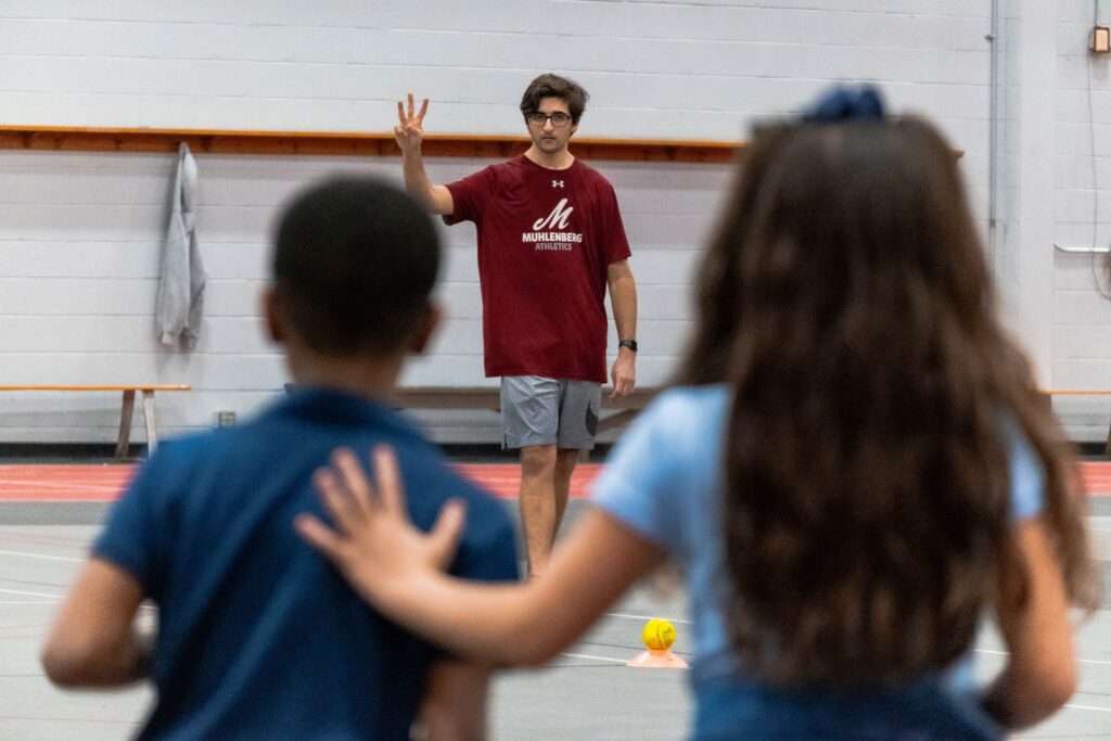 A college student holds up three fingers as elementary school students wait their turn to run in a gym