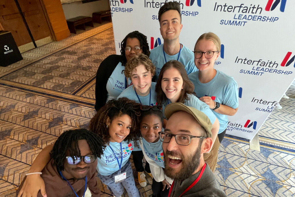 A selfie of college students and a college staff member in front of a sign that says Interfaith Leadership Summit