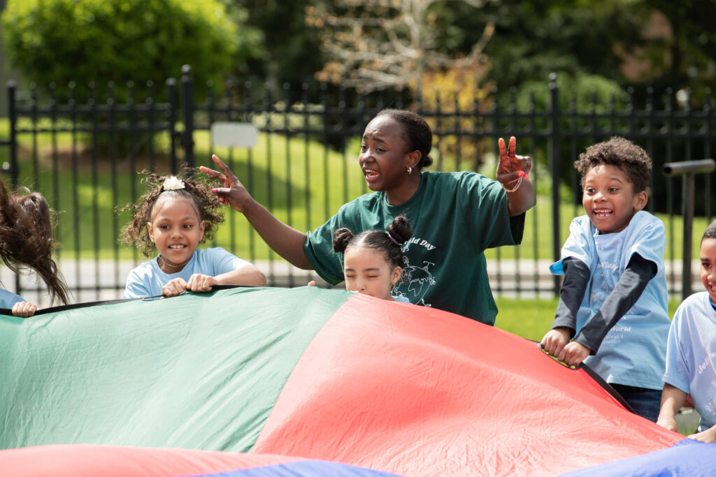 A college student stands with elementary school students who are using a colorful parachute