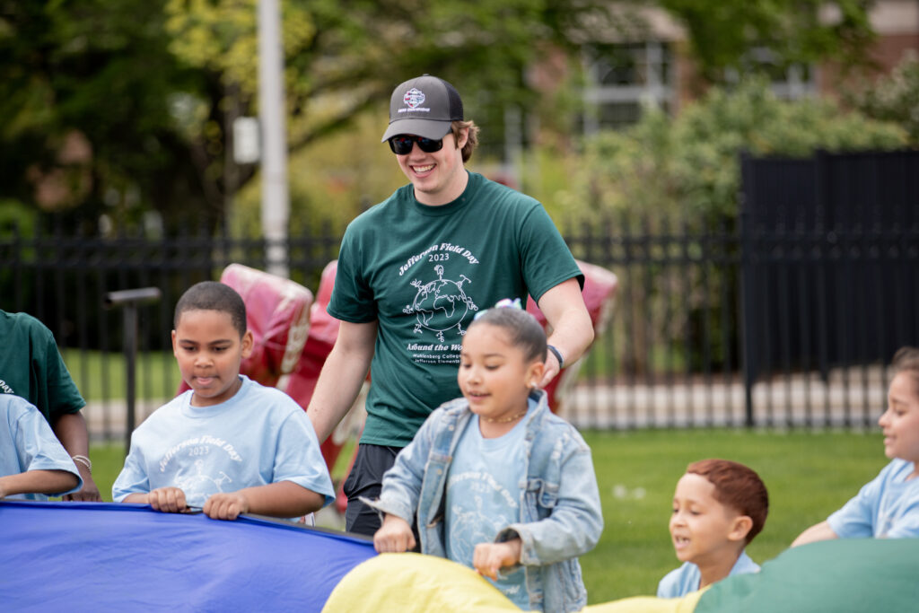 A college student in a hat and sunglasses helps young children playing with a parachute