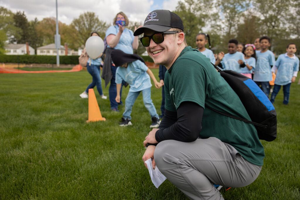 A college student squats in the grass and smiles while watching young children play a game