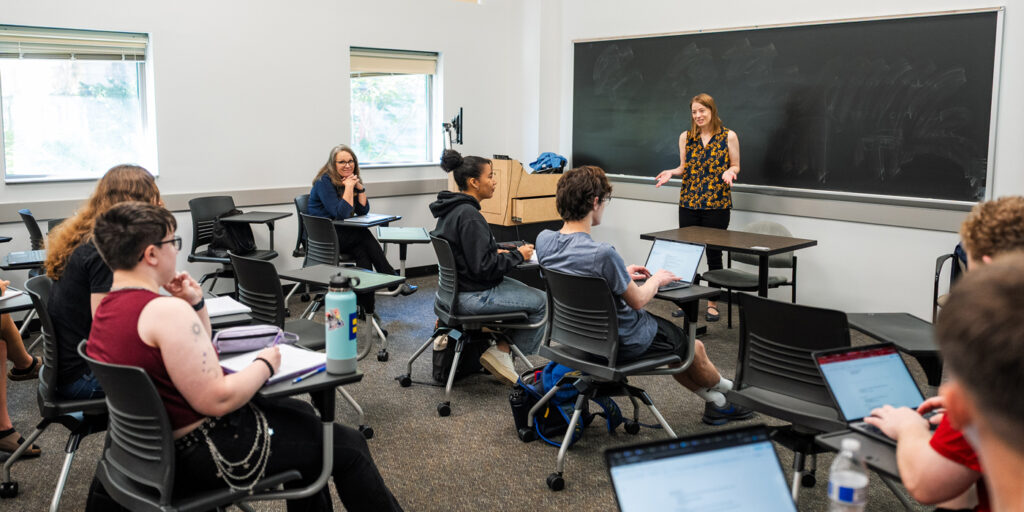 A college professor with red hair stands in front of a blackboard and speaks to a classroom of students