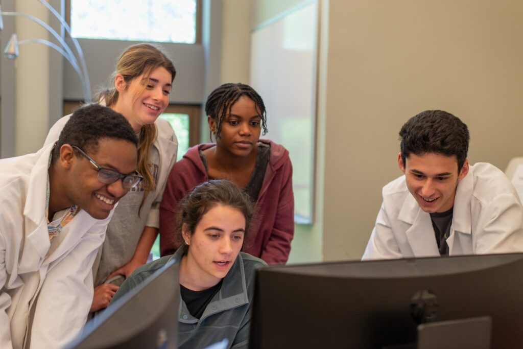 Five students in a lab, some of whom wear lab coats, look at a computer screen.