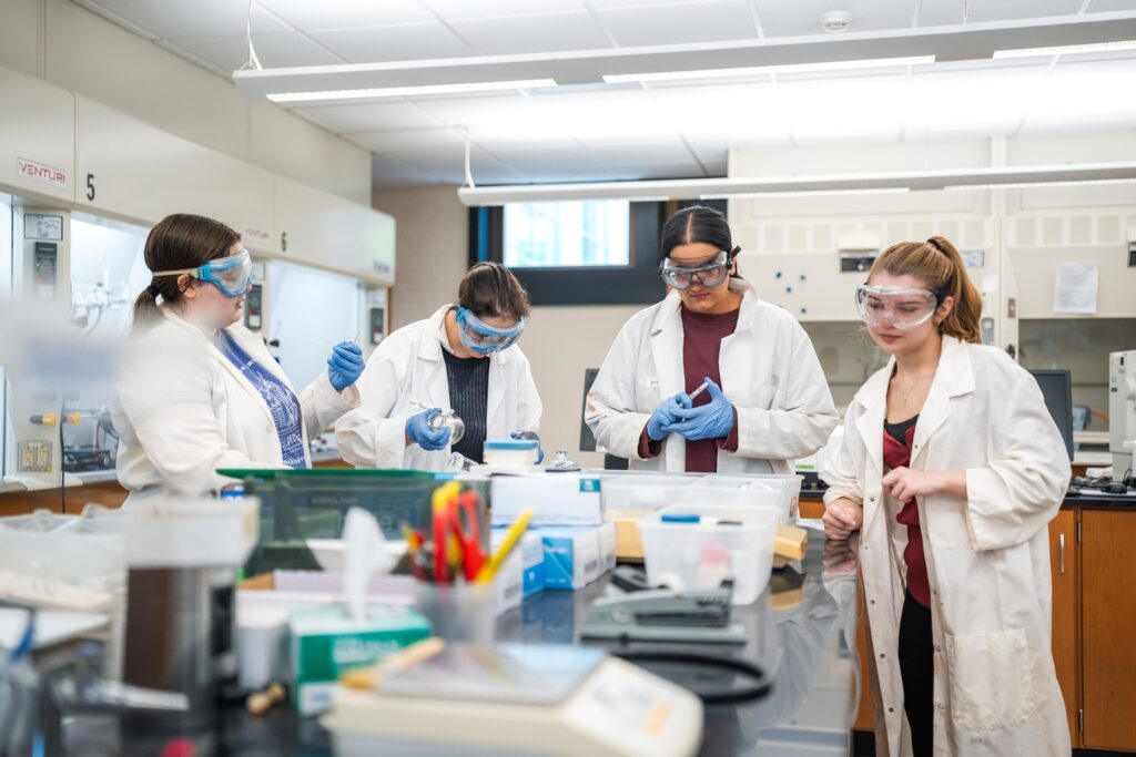 Four students wearing white lab coats and gloves in a lab conduct research.