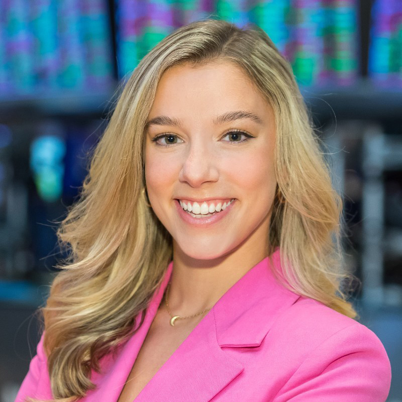 A woman with long blond hair wearing a pink blazer smiles in front of a trading floor