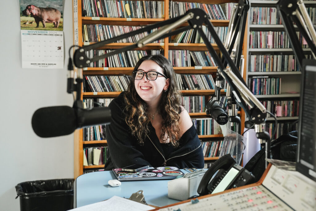 A college student with long dark hair and glasses smiles in a radio studio with CDs behind her