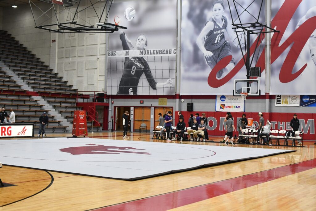 Wide view of a gymnasium set up for a wrestling meet, with a large white wrestling mat centered on the hardwood floor. Team benches and athletes line the far side of the mat, while bleachers rise on the left. Large wall banners show Muhlenberg College athletes, and a basketball hoop hangs above the court.