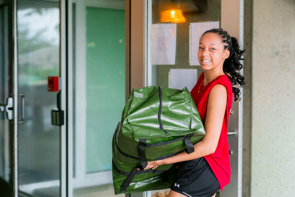A Muhlenberg College student in a red shirt carries a bag and smiles at the camera during move-in