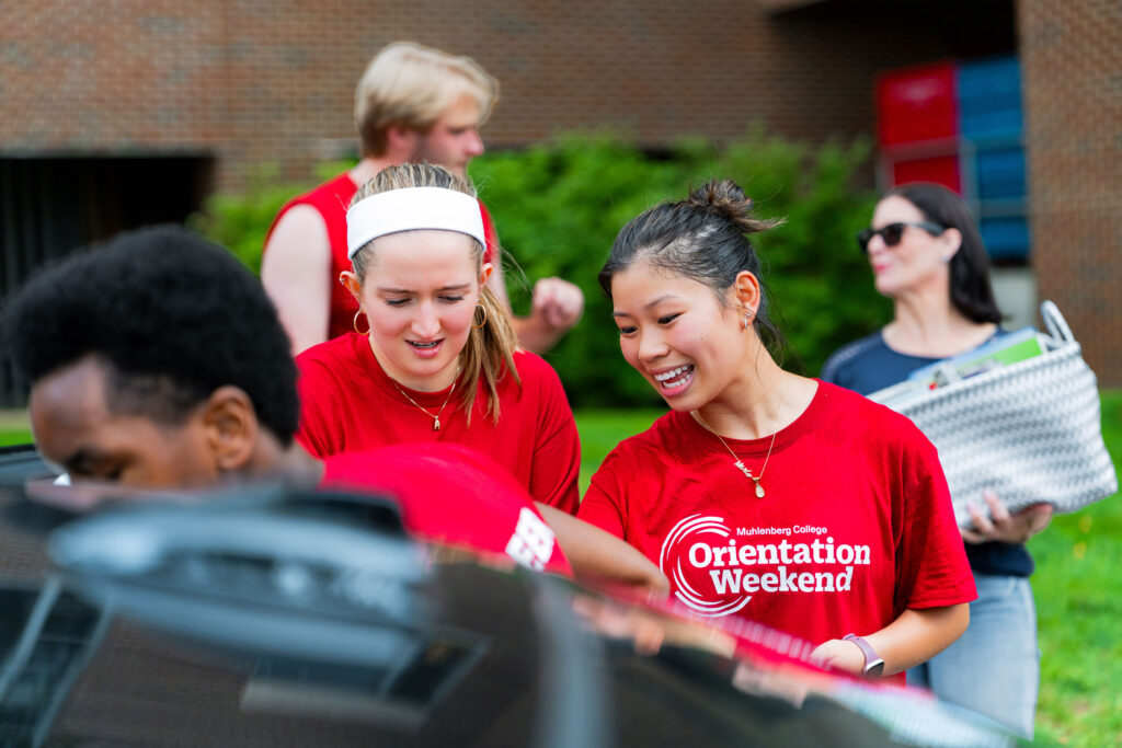 Muhlenberg College students in red shirts gather around a car to carry belongings during move-in