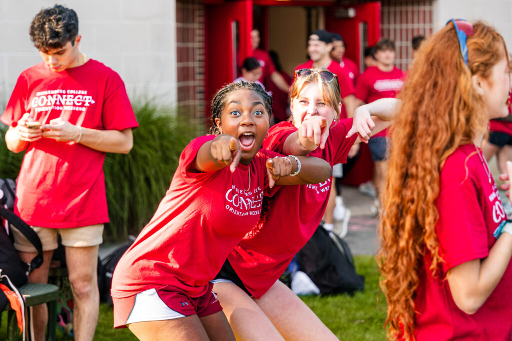 Two college students in red orientation T-shirts point at the camera during move-in
