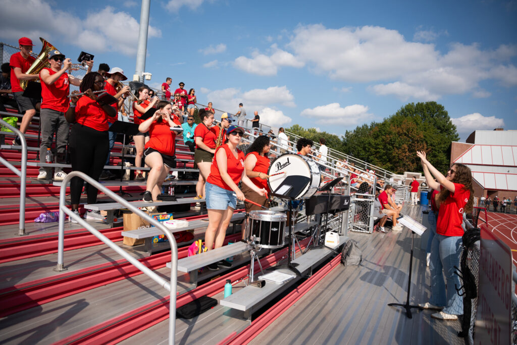 A college pep band wearing matching red T-shirts performs in the stands during a football game
