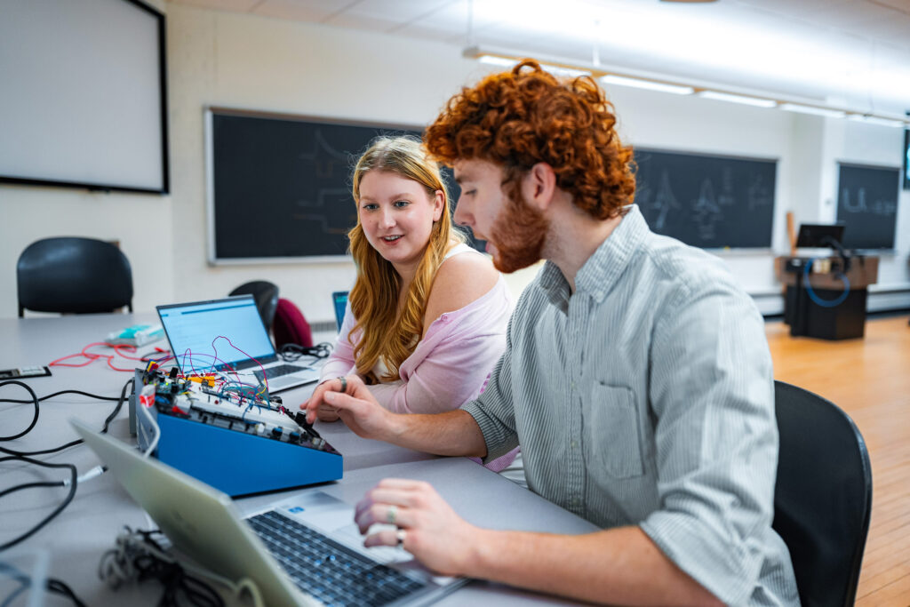 A pair of college students work together in front of a circuit board in a physics lab.