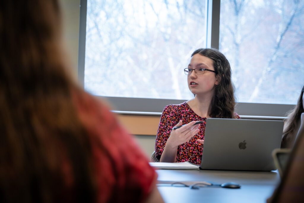 A college students speaks up while seated at a desk in a classroom.