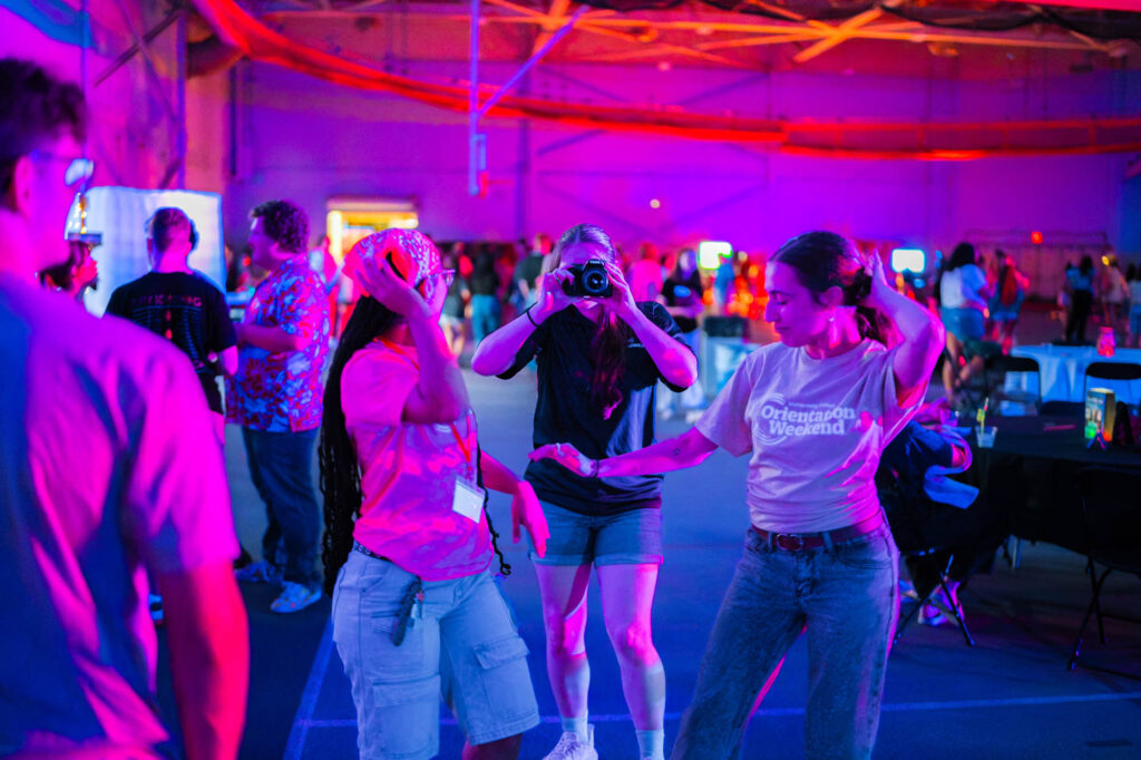 A Muhlenberg College student uses a camera to take a photo while two students dance next to her in the field house