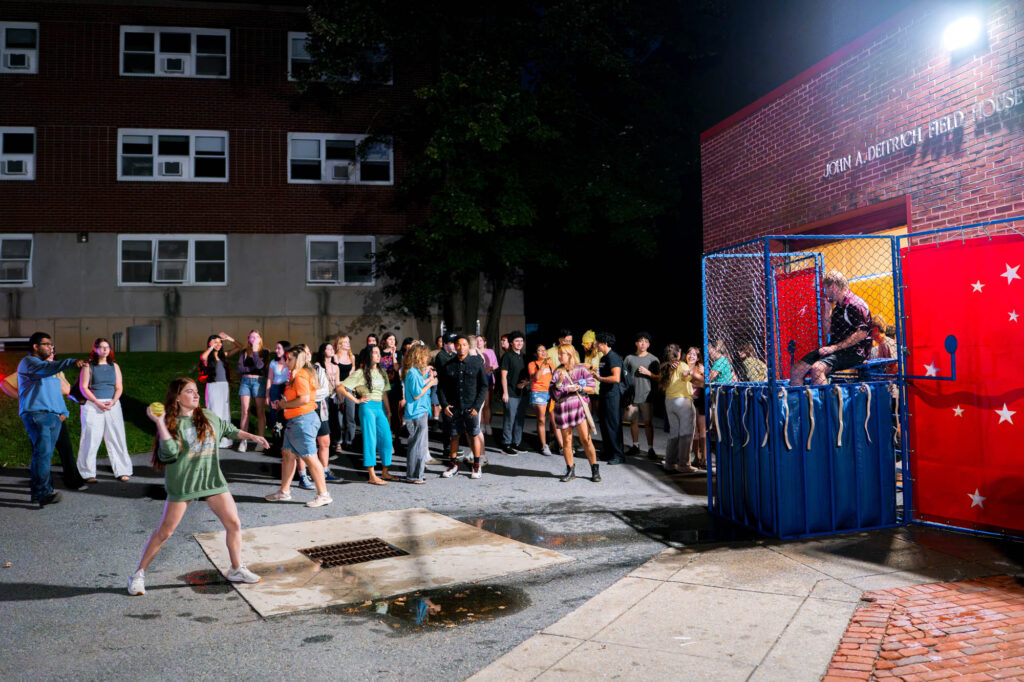 A Muhlenberg College student throws a ball at a dunk tank outside while other students watch