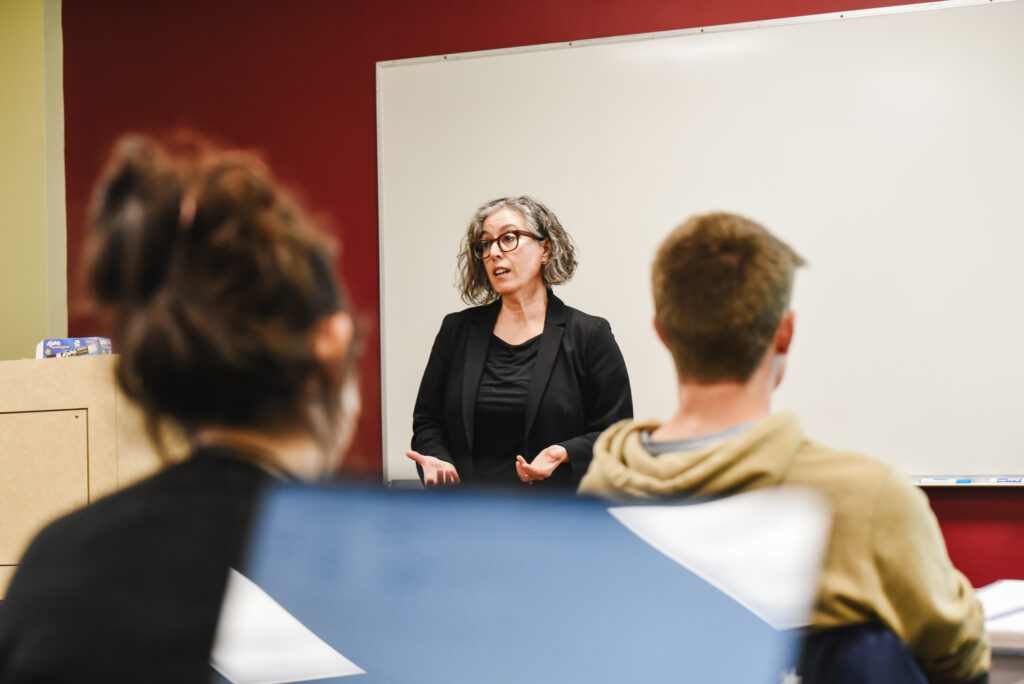 A college professor wearing glasses and a black blazer teaches a class