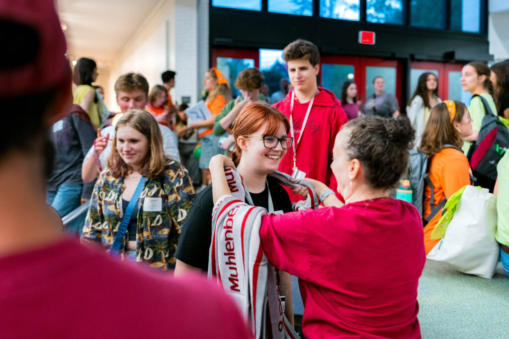 A Muhlenberg College alum drapes a scarf over a current student with red hair and glasses