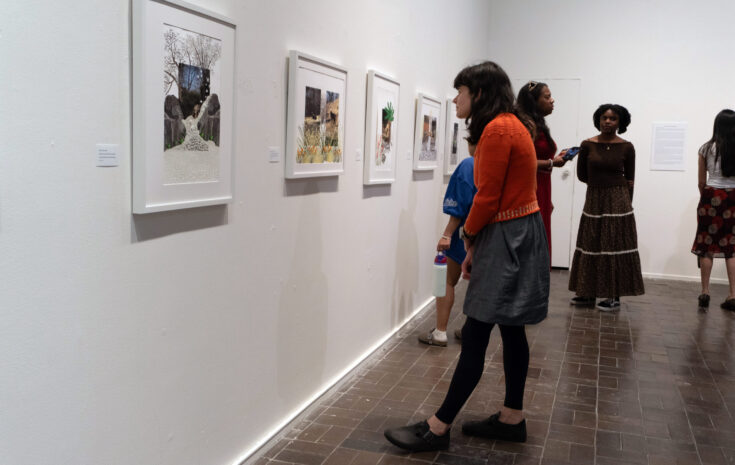 A woman in an orange sweater looks at a piece of art hanging on a gallery wall