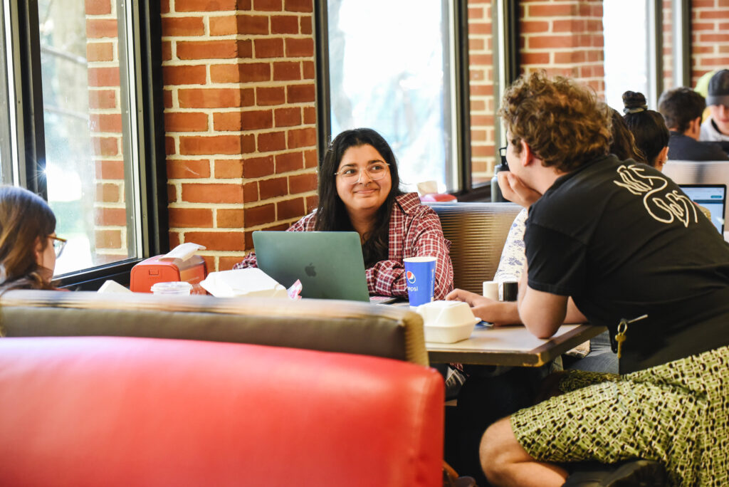 A college student smiles at another student while sitting in a booth