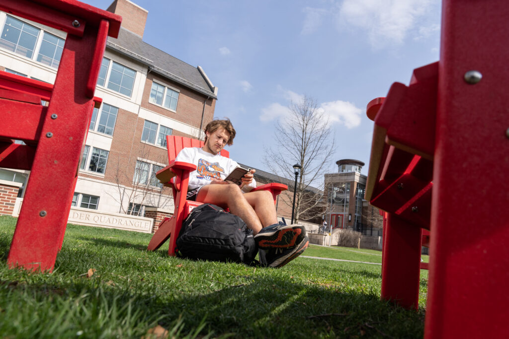 A Muhlenberg student reads a book outdoors in a red Adirondack chair.