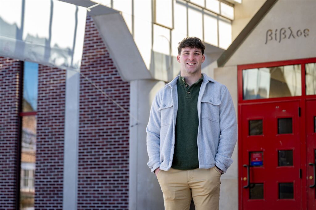 A person stands smiling in front of a brick building with red doors and glass windows, wearing a light gray jacket and tan pants, conveying a welcoming, casual tone.