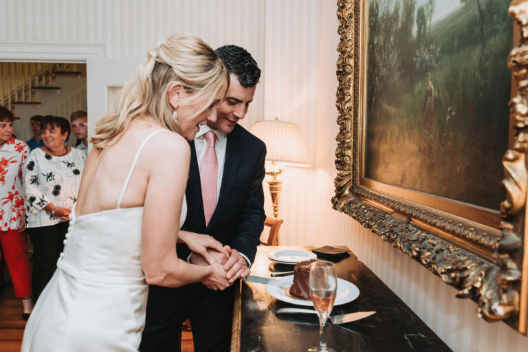 A bride and groom cut a piece of cake next to an oil painting