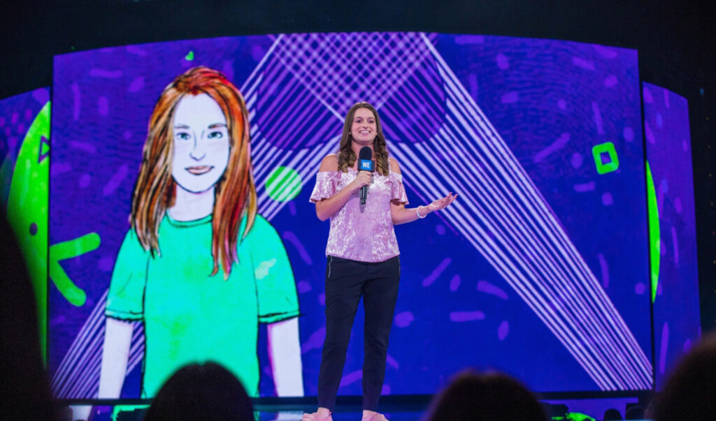 A Muhlenberg alum stands on a stage with a microphone, child artwork projected on a screen behind.