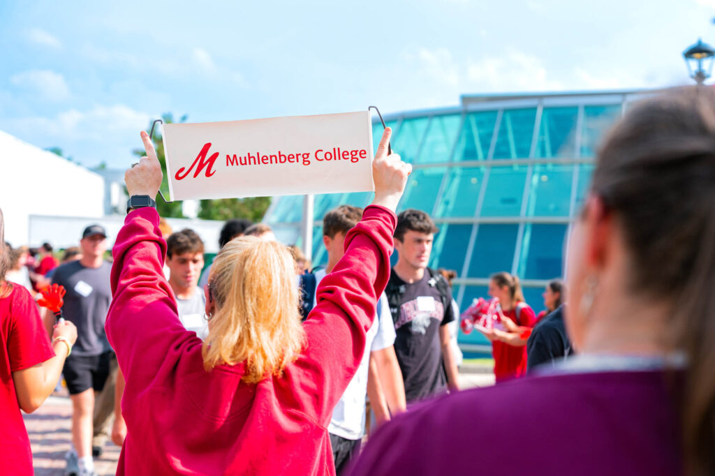 A woman holds up a sign that says Muhlenberg College while new students walk by