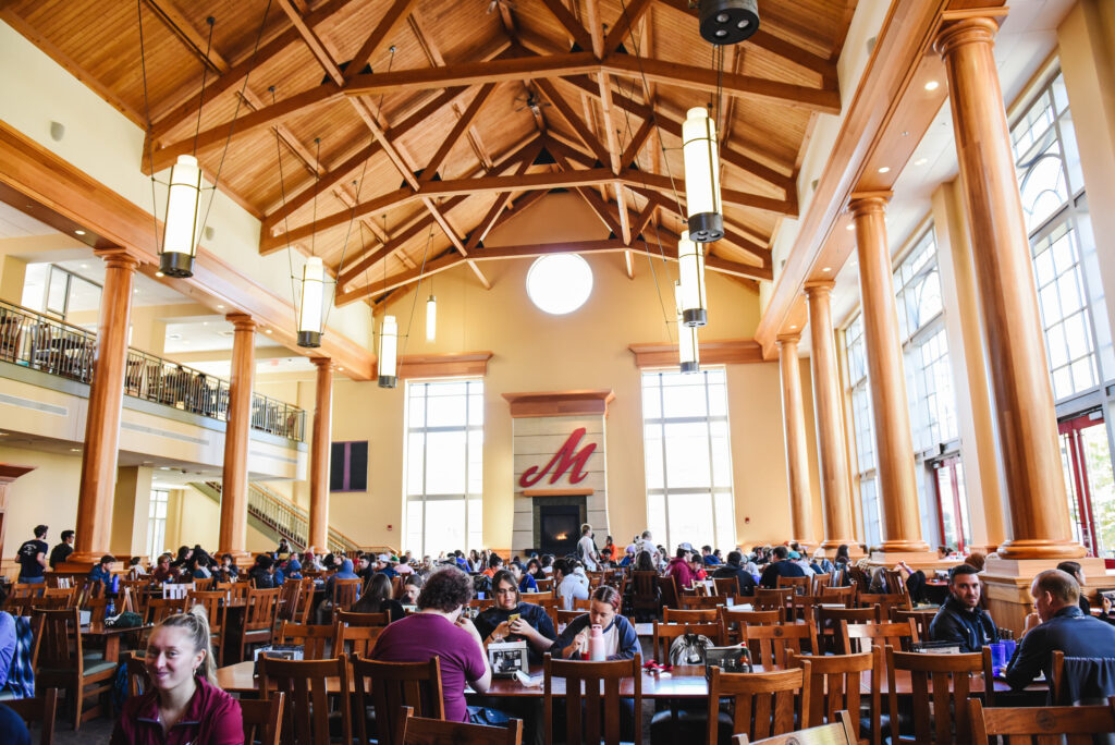 A college dining hall with high wooden ceilings and a fireplace with a large red M over it
