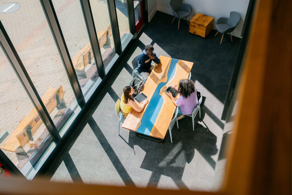 An aerial photo of three college students working at a wooden table next to a large window