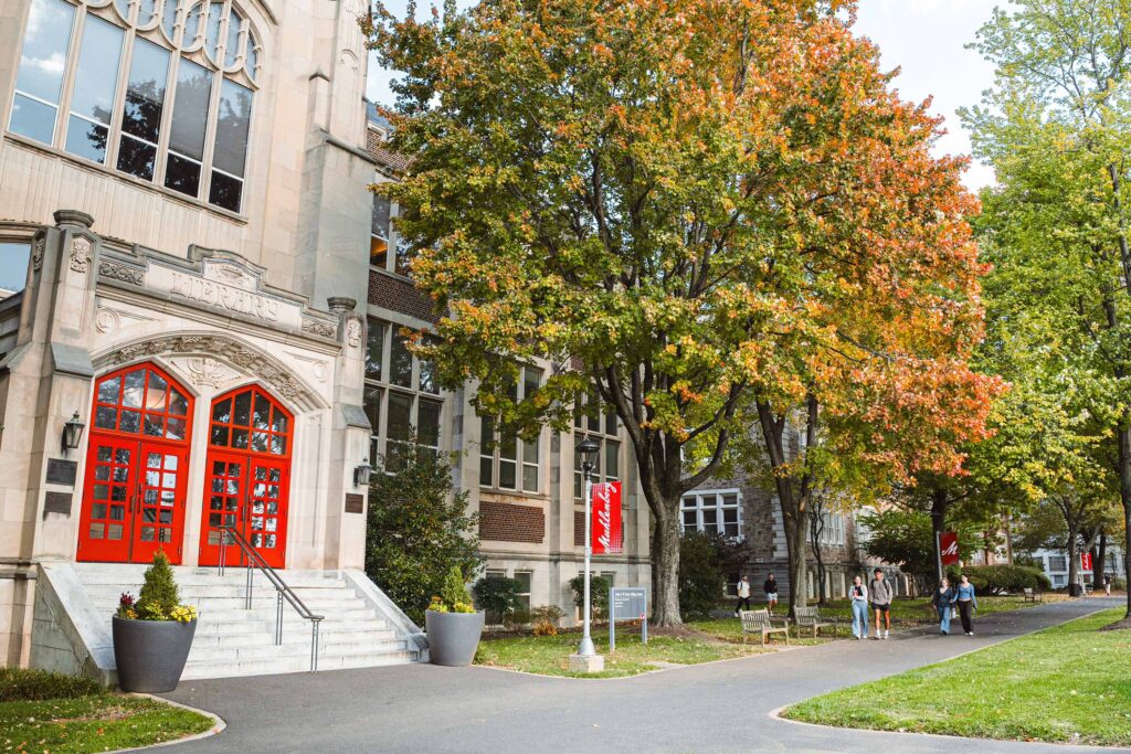 Students walk outside an academic building with red doors near a tree with orange leaves