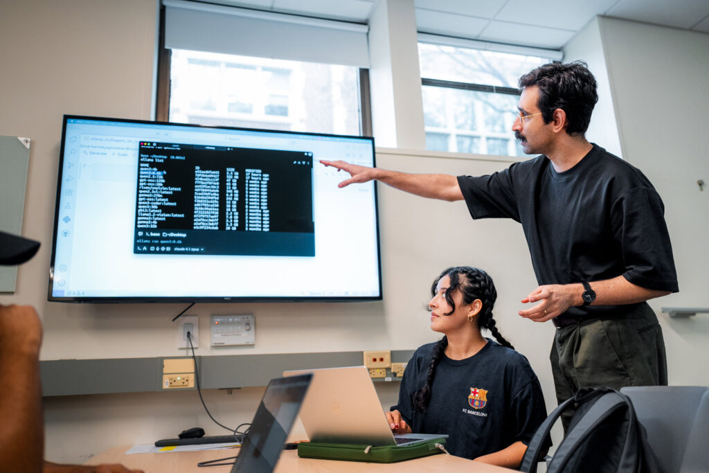 A college professor gestures at a screen while a college student on a laptop looks on