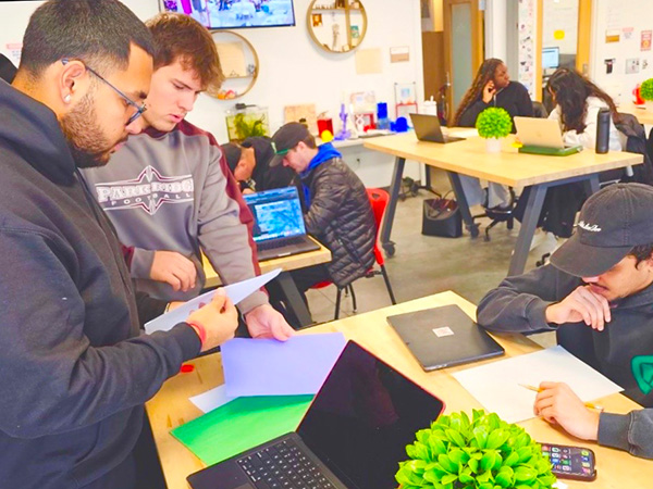 Students in a classroom collaborating around a table with laptops and paperwork